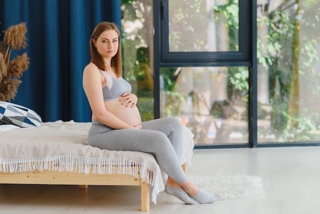 O conceito de gravidez e espera - uma mulher grávida feliz segura o estômago com as mãos, sentada em uma cama