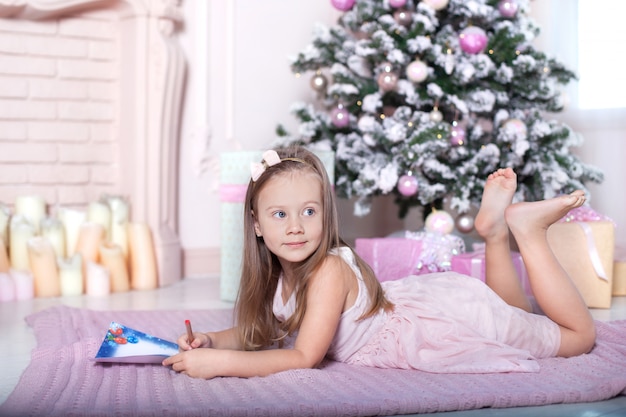 O conceito de natal, férias e infância. menina criança escreve uma carta para o papai noel em um quarto infantil perto da árvore de natal. véspera de natal. tradições familiares. inverno