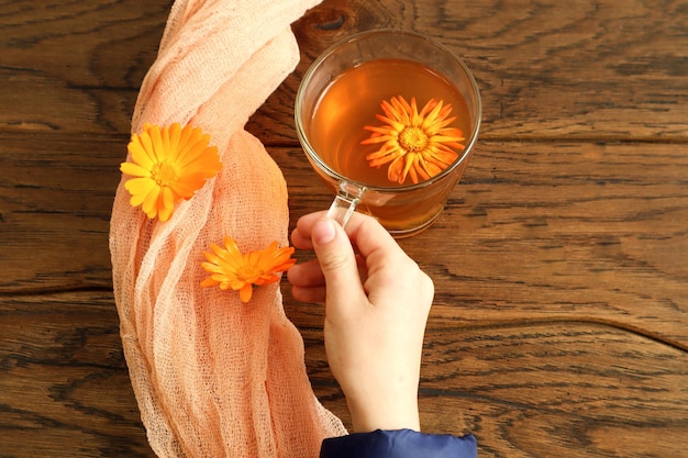 O conceito de tomar chás de ervas medicinais. Copo de vidro com chá na mão, próximo a um guardanapo com flores de calêndula, vista de cima, local para texto