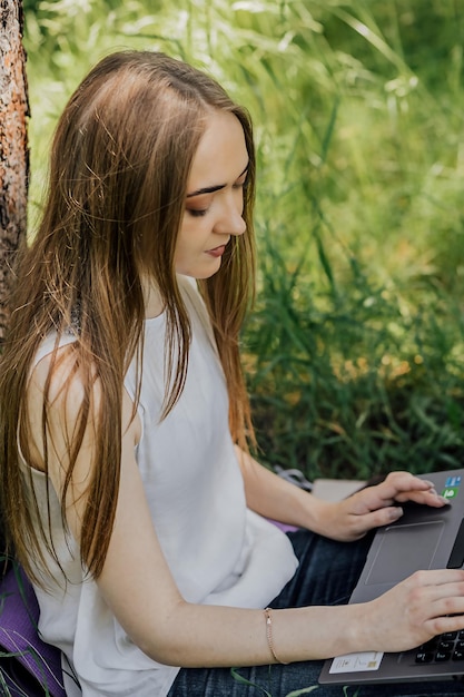 O conceito de trabalho remoto Uma jovem trabalha com um laptop ao ar livre no parque sentado no gramado Trabalhar como freelancer A garota faz cursos em um laptop e sorri