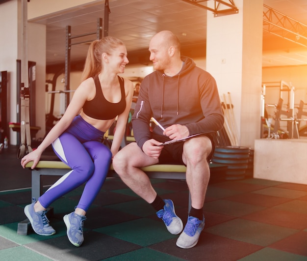 Foto o conceito de treinamento pessoal. notas do instrutor no caderno resulta em treinamento desportivo mulher loira sentada em um banco. casal sorridente de esportes