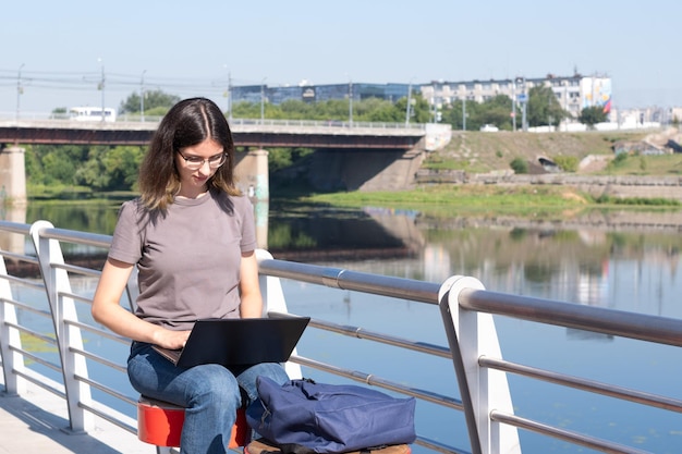 O conceito de um freelancer Uma bela estudante morena com óculos está usando um laptop na rua