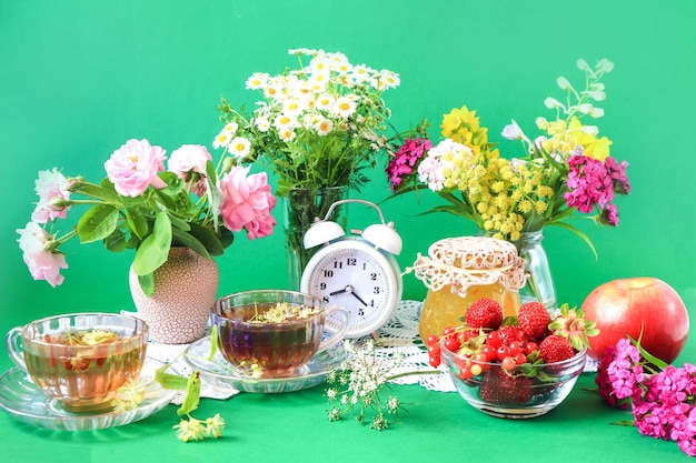 O conceito de uma boa manhã de verão Xícaras de chá entre buquês de flores silvestres morangos em uma xícara potes de maçãs compota despertador fundo verde vista lateral