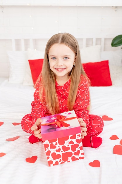 o conceito do Dia dos Namorados uma criança bonita está sentada na cama em casa de pijama vermelho e segurando um presente em suas mãos e sorrindo ou rindo de felicidade parabenizando pelo feriado