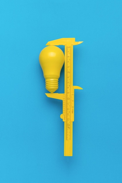 O conceito mínimo da ideia Um paquímetro amarelo medindo uma lâmpada elétrica amarela em um fundo azul Precisão e energia
