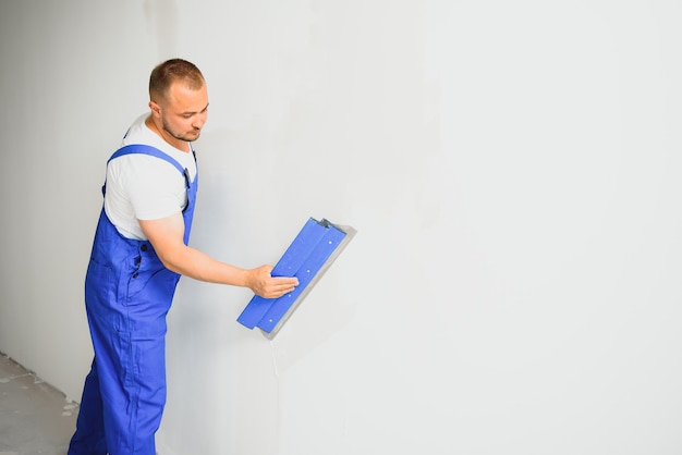 O construtor corrige cuidadosamente as irregularidades da parede com uma talocha. Construtor com roupa de trabalho contra uma parede cinza. Estucador de fotos no trabalho.