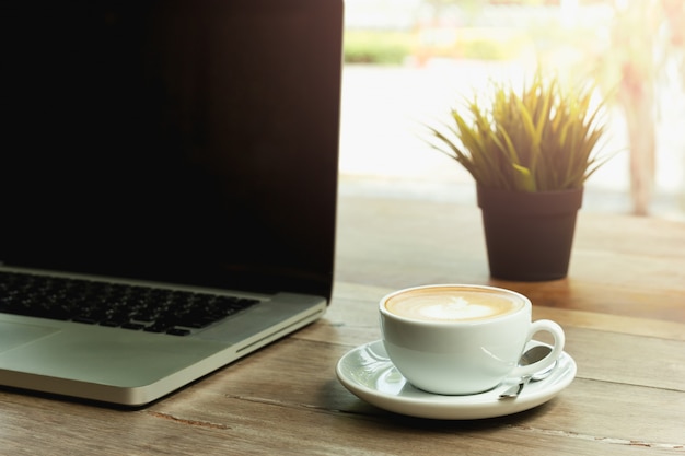 O copo de café com o laptop na tabela e na manhã de madeira ilumina-se.