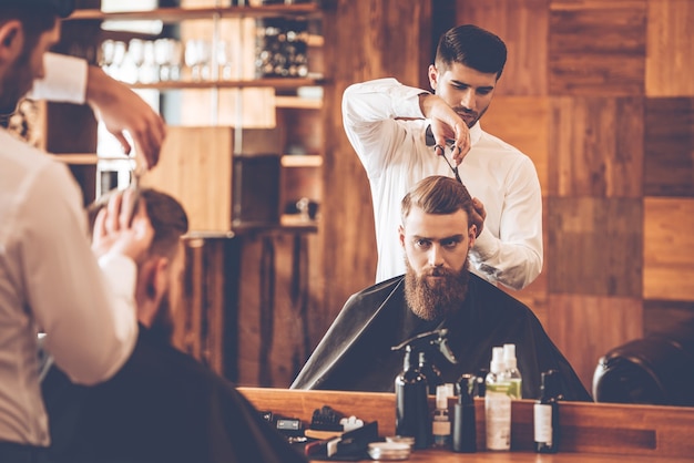 Foto o corte de cabelo deve ser perfeito. jovem barbudo cortando cabelo de cabeleireiro enquanto está sentado na cadeira de uma barbearia em frente ao espelho