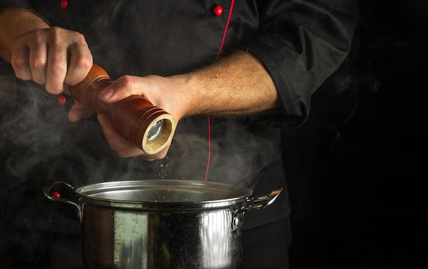 O cozinheiro adiciona pimenta moída a uma panela de comida fervente Conceito de cozinha de restaurante com espaço publicitário em fundo preto