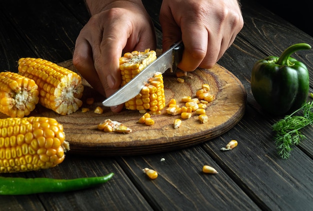 O cozinheiro corta o milho com uma faca em uma tábua O milho é um excelente café da manhã ou almoço dietético preparado pelas mãos do chef