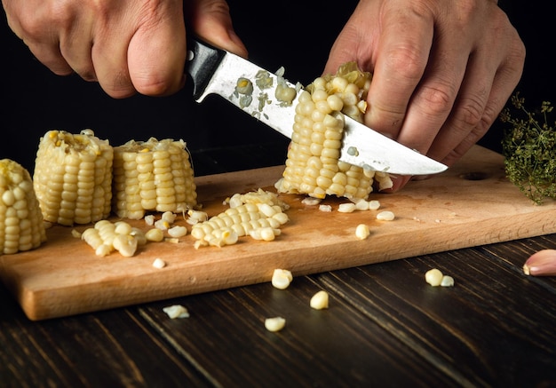 O cozinheiro corta os grãos de milho cozido com uma faca O milho é um excelente café da manhã ou almoço dietético pelas mãos do chef