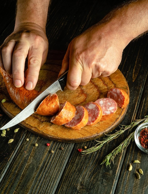 O cozinheiro corta salsicha de carne de porco com uma faca em uma tábua de corte de madeira O conceito de preparar deliciosas sanduíches para o almoço para um lanche