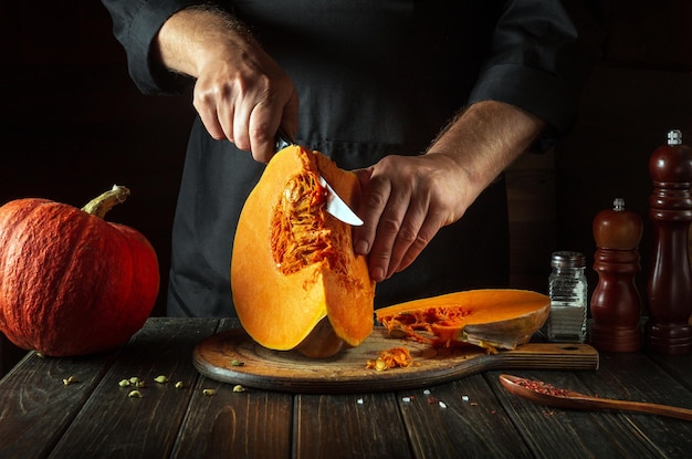 O cozinheiro corta uma abóbora laranja em fatias com uma faca em uma tábua de corte de madeira