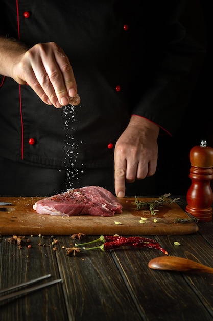 Foto o cozinheiro espalha sal no bife de carne para cozinhar para o almoço de churrasco ambiente de trabalho na mesa da cozinha com rosmarino e alho perfumados