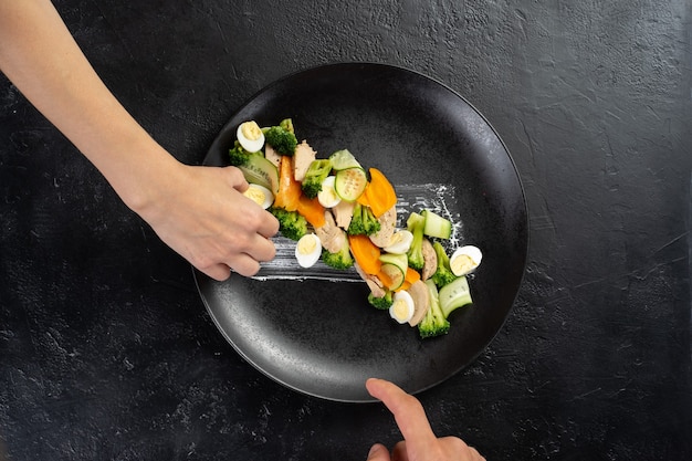 O cozinheiro prepara uma salada de legumes com couve-flor, cenoura, pepino, frango e ovos de codorna em um prato preto com as mãos.