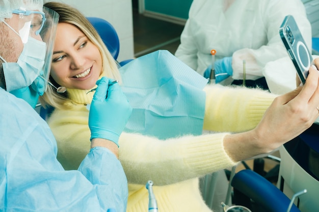 O dentista com máscara protetora senta-se ao lado do paciente e tira uma foto após o trabalho.