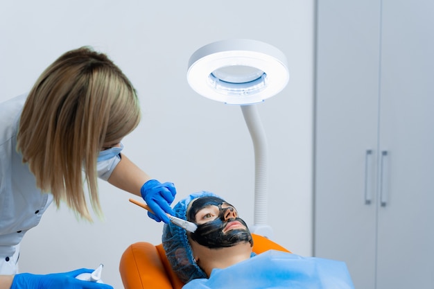 O dermatologista passa máscara preta no rosto para fotorejuvenescimento a laser e peeling de carbono. Dermatologia e cosmetologia. Usando laser cirúrgico.