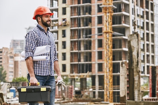 O dia de trabalho está começando, jovem construtor alegre de capacete vermelho segurando luvas protetoras de caixa de ferramentas e