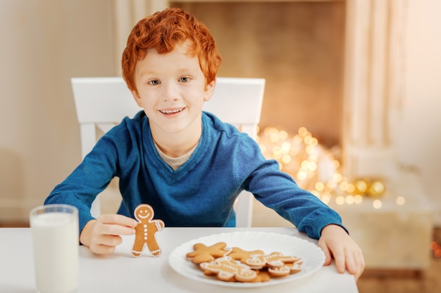 O dia mais emocionante. Adorável menino de cabelos cacheados, sentado à mesa e sorrindo alegremente enquanto aprecia seu delicioso café da manhã em uma manhã de Natal.