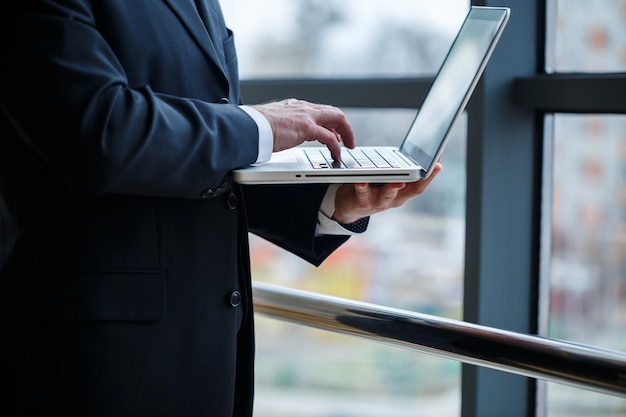 O diretor executivo sorridente e feliz pensa no desenvolvimento de sua carreira de sucesso enquanto está de pé com um laptop em seu escritório perto do fundo de uma janela com espaço de cópia