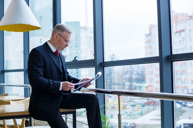 O diretor-gerente homem encostou-se em uma mesa de madeira em seu escritório perto de uma grande janela e analisa um plano de desenvolvimento de negócios bem-sucedido.