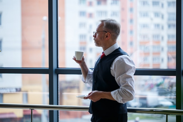 O diretor-gerente sorridente e feliz pensa no desenvolvimento de sua carreira de sucesso enquanto está em pé com uma xícara de café na mão em seu escritório, perto do fundo de uma janela com espaço de cópia