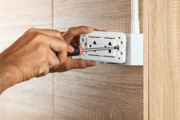 O eletricista está usando uma chave de fenda para instalar a tomada elétrica em uma caixa de tomada de plástico em uma parede de madeira.