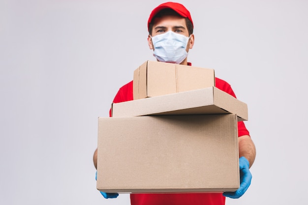 O empregado do homem de entrega nas luvas uniformes da máscara protetora do t-shirt vermelho do espaço em branco da tampa mantém a caixa de papelão vazia isolada na parede branca. serviço conceito de vírus de coronavírus pandêmico de quarentena 2019-ncov.