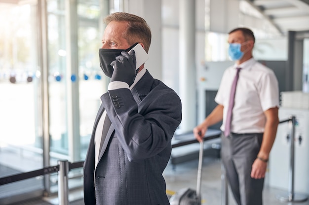 O empresário com luvas de proteção e máscara está falando no celular enquanto o assistente carrega a bagagem no aeroporto