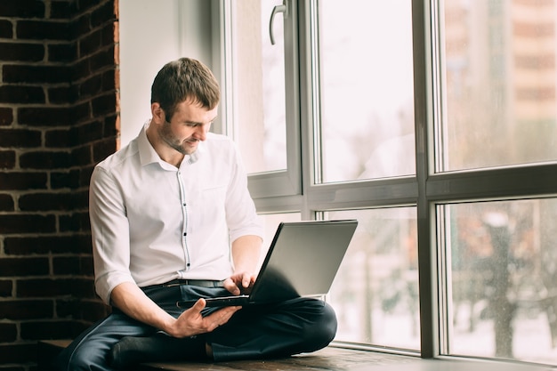 O empresário se senta no parapeito da janela em pose de lótus e trabalha no laptop em seu gabinete