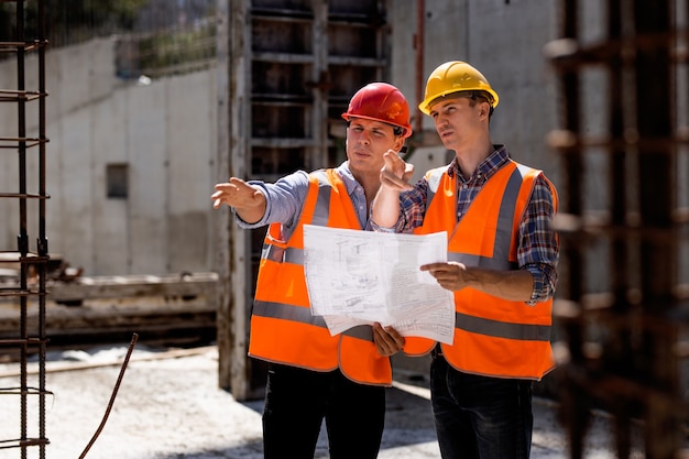 O engenheiro civil e gerente de construção com coletes laranja e capacetes rígidos exploram a documentação da construção no canteiro de obras perto das estruturas de aço. .