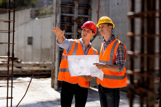 O engenheiro civil e gerente de construção com coletes laranja e capacetes rígidos exploram a documentação da construção no canteiro de obras perto das estruturas de aço. .