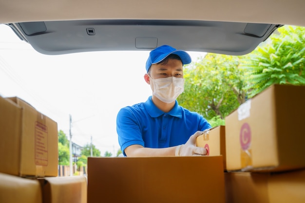 O entregador asiático presta serviços de correio para trabalhar com caixas de papelão em uma van durante a pandemia de Coronavirus (COVID-19), correio usando máscara médica e luvas de látex para segurança.