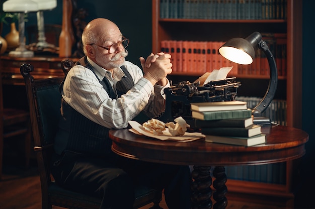 O escritor idoso pensa na máquina de escrever vintage no escritório em casa. velho de óculos escrevendo romance de literatura em uma sala com fumaça