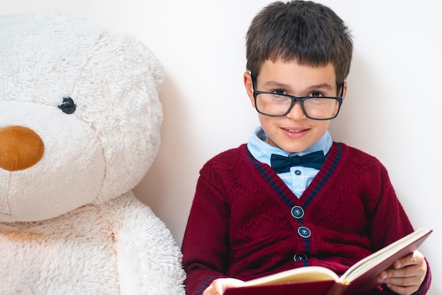 O estudante de suéter e óculos está sentado ao lado de um grande ursinho de pelúcia com um livro aberto nas mãos