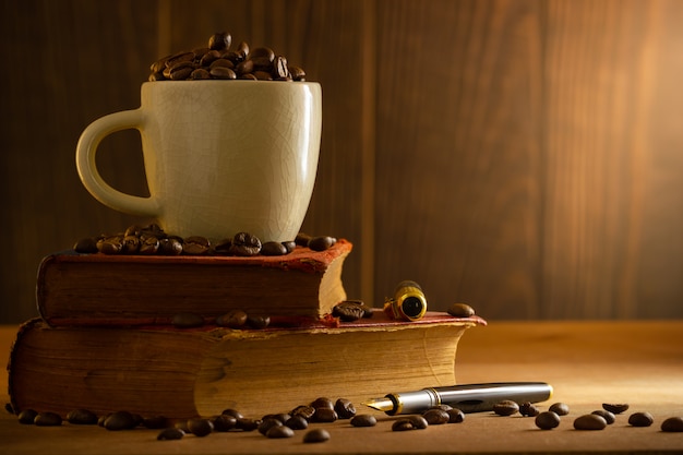 O feijão de café no copo branco e o vintage registram o empilhamento na tabela de madeira na luz da manhã.