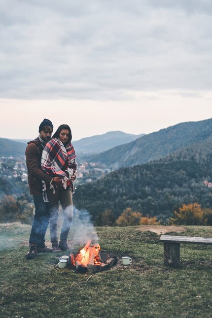 O fogo os mantém aquecidos. Jovem bonito abraçando sua namorada atraente enquanto ficava perto da fogueira ao ar livre