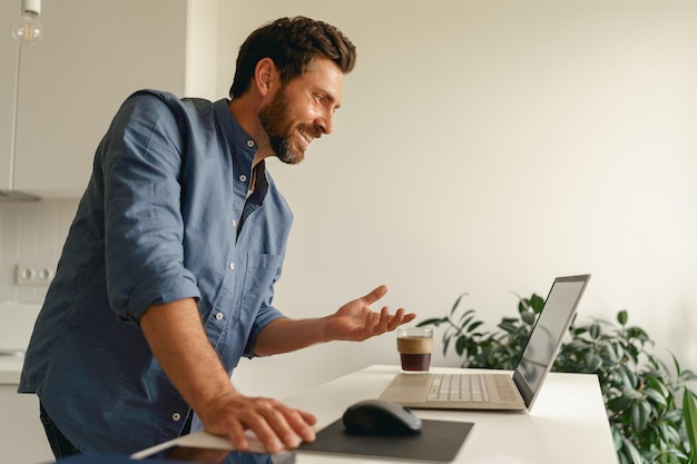 O freelancer masculino tem uma videoconferência do laptop durante o trabalho à distância da cozinha doméstica