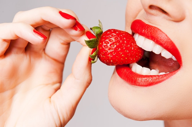 O frescor vem em vermelho. Close de uma linda mulher segurando um morango na mão e provando-o em pé contra um fundo cinza