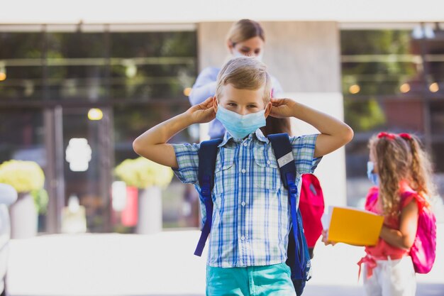 O garoto do ensino fundamental usando uma máscara protetora O estudante com uma máscara médica fica na frente da escola