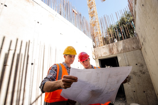 Foto o gerente de construção e o engenheiro vestidos com roupas de trabalho e capacetes exploram a documentação do projeto no canteiro de obras. .