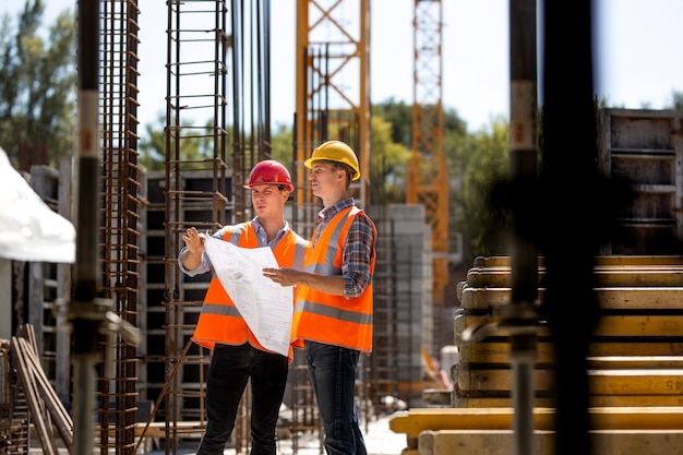 O gerente e engenheiro de construção vestido com coletes laranja e capacetes rígidos exploram a documentação da construção no canteiro de obras próximo às estruturas de aço. .