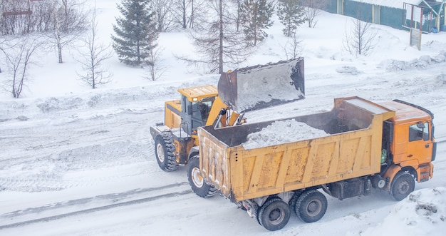 O grande trator amarelo limpa a neve da estrada e carrega-a no caminhão.