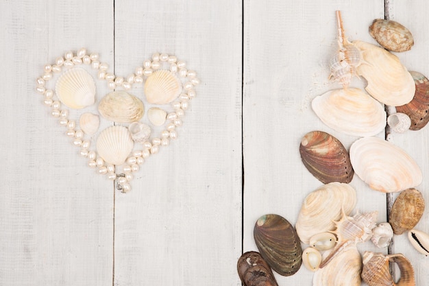 Foto o grupo de conchas do mar e pérolas dispostas em forma de coração sobre fundo branco de madeira