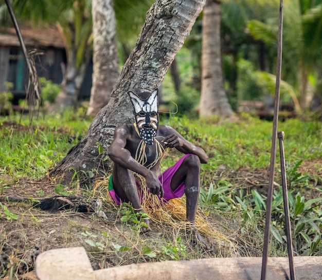 O guerreiro da tribo Asmat com uma máscara de combate está sentado perto de uma árvore.