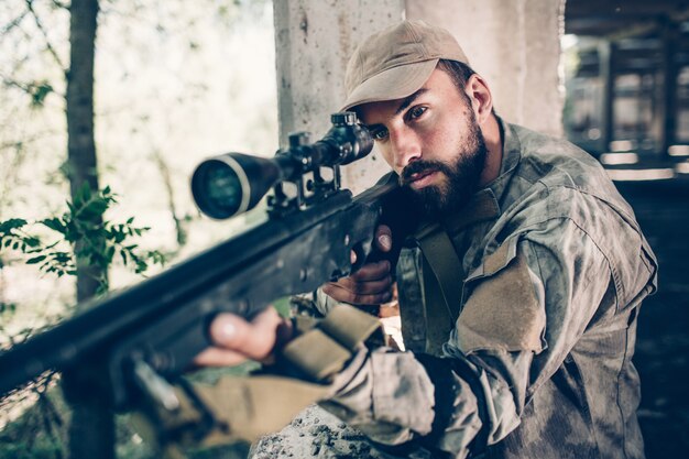 O guerreiro sério e corajoso está parado perto da saída do hangar e olhando através da lente. Ele está mirando usando rifle. Cara barbudo está concentrado. Ele está esperando.