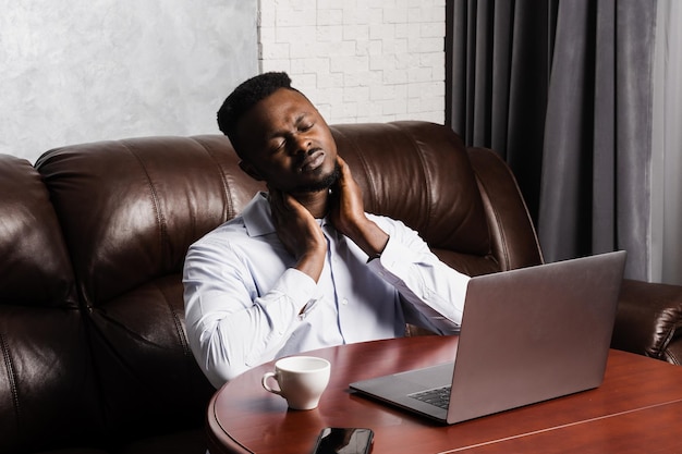 O homem africano sente dor no pescoço depois de trabalhar por longas horas com o laptop online no escritório A osteocondrose da coluna cervical é uma síndrome radicular do homem afro-americano