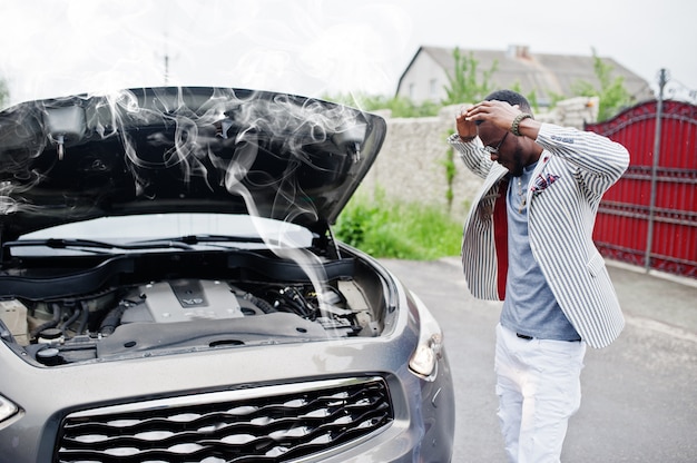 O homem afro-americano elegante e rico está na frente de um carro quebrado suv precisa de assistência, olhando sob o capô aberto com fumaça.