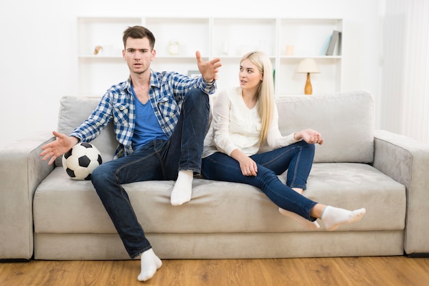 O homem animado assiste a uma bola de futebol perto de uma mulher no sofá