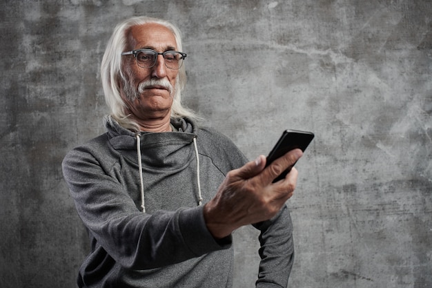 O homem aposentado caucasiano grisalho envelhecido parece surpreendido na tela do telefone móvel. Avô em copos com bigode, dominando a tecnologia moderna.